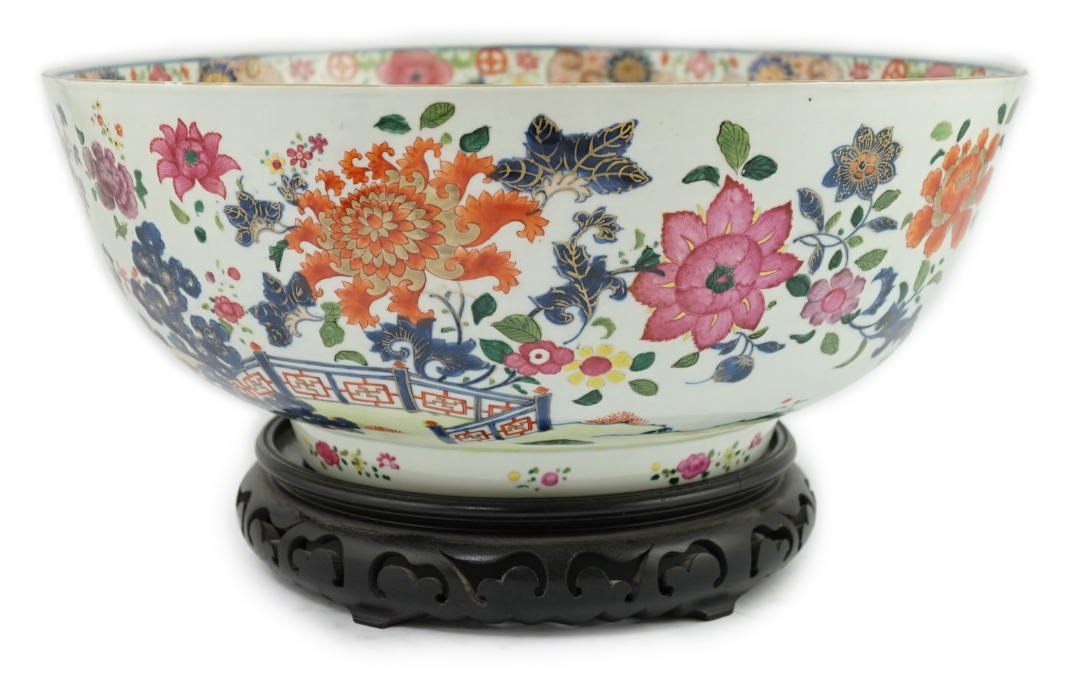 A large Chinese famille rose punch bowl, Qianlong period, the exterior painted with flowers in a fenced garden, the interior with a flower bouquet within a garland of flowers, 40.5cm diameter, wood stand. Condition - som